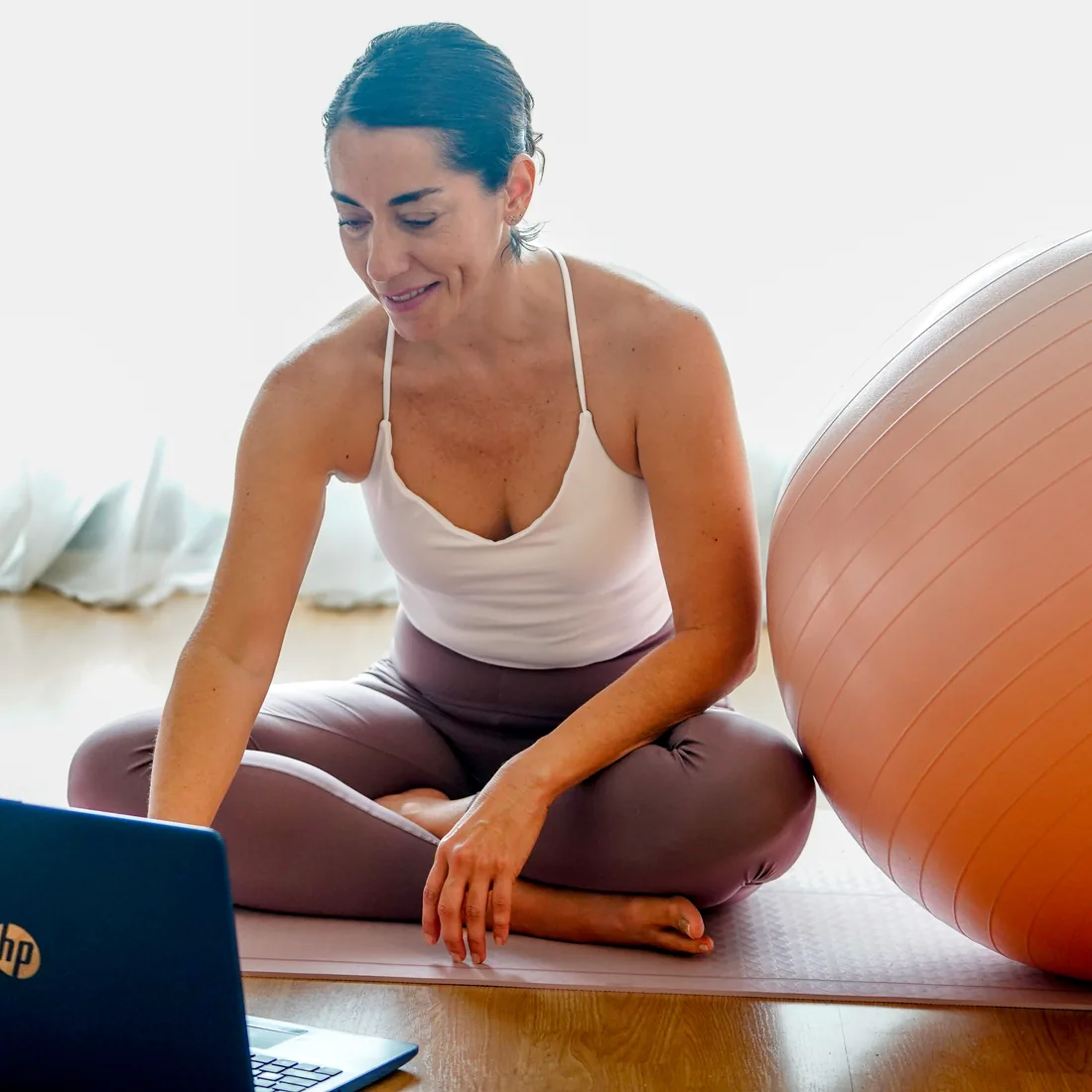Belén sentada en la colchoneta junto a un fitball, sonriendo mientras prepara la sesión frente al portátil